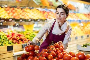 Female employee informs about arrival of ripe tomato, advertises product. Assistant seller in apron, forms display case in vegetable store. Local and imported farm vegetables and fruits