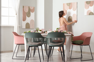 Pretty young woman hanging painting on wall in light dining room with elegant table setting