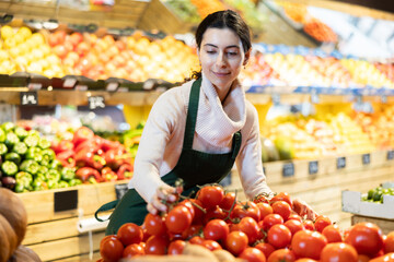 Young woman seller in apron puts tomatoes on display in vegetable shop