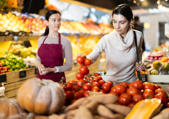 While shopping in vegetable shop, woman customer takes ripe tomato from box on display case. Sales assistant monitors order in shop, standing behind customer, offer consulting to client