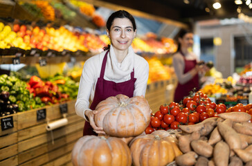 Woman seller in an apron stands in the vegetable section of a supermarket and offers a seasonal pumpkin. Grocery store worker checks pumpkin quality and invites to shop
