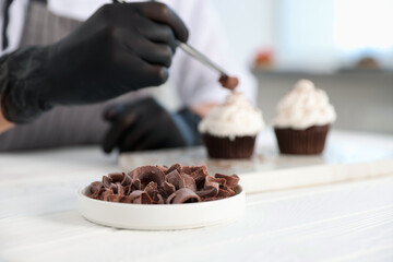 Confectioner decorating tasty cupcake with chocolate shaving at white wooden table in kitchen, selective focus
