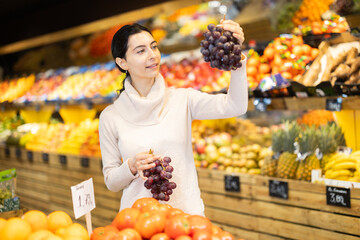 Near shelves and display cases in sales area of store, woman customer takes ripe grape out of box. Wide range of agricultural products. Self-service, comfortable shopping.