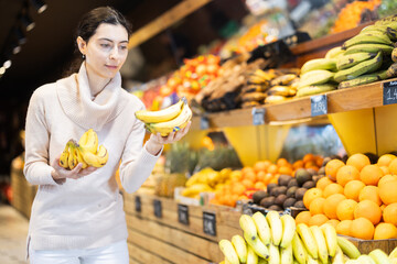 Young woman buyer choosing bunch of fresh bananas in vegetable shop