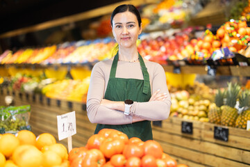 Positive middle-aged female seller standing by tomato stand in fruit and vegetable market