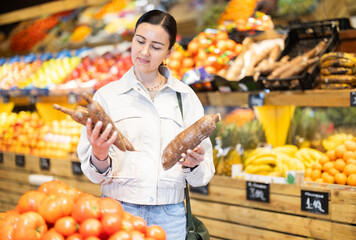 Positive middle-aged woman choosing cassavas standing at counter in large vegetable market
