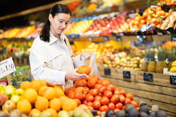 Positive middle-aged woman choosing tomatoes standing at counter in large vegetable market