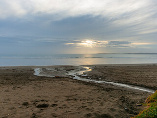 Small stream leads across beach to waters edge as sun approaches horizon through overcast sky