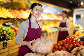Adult woman seller in apron puts pumpkin on display in vegetable shop