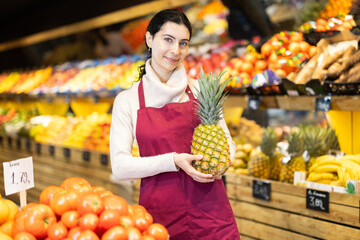 Vegetable shop assistant greets customers with goods in hands. Woman seller in sales area with pineapple in hands. Employee demonstrates supply of fruit. Local and imported farm vegetables and fruits