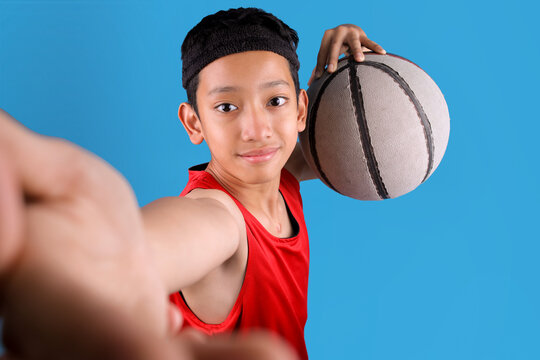 Portrait of smiling Asian man holding ball and taking selfie while playing basketball on blue background