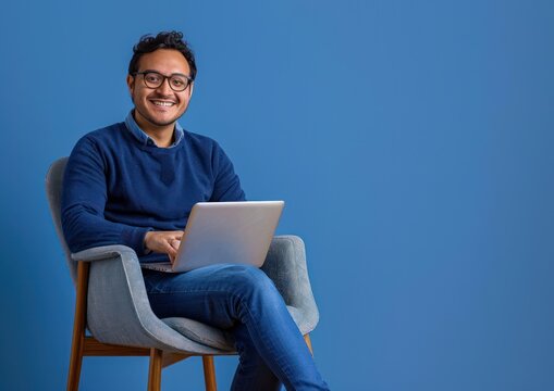 smiling man wearing glasses and blue sweater sitting comfortably on a gray chair with a laptop on his lap against a solid blue background