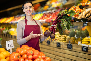 Polite middle-aged female seller offering beetroot standing by counter in large grocery store