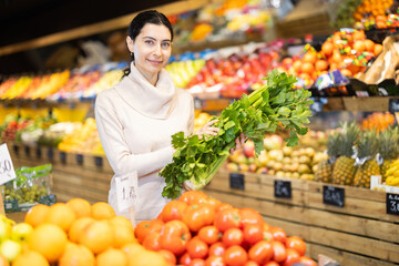 In vegetable store, young woman customer buy ripe celery. Vegetables and fruits from different parts of world, products from local farms.
