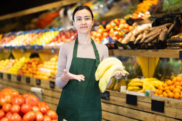Polite middle-aged female seller offering bananas standing by counter in large grocery store