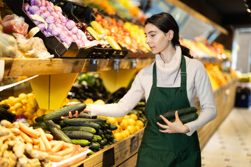 Young woman seller in apron puts fresh cucumbers on display in vegetable shop