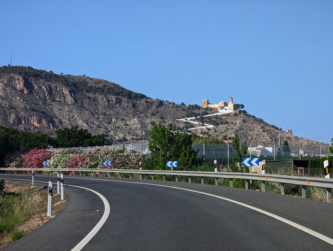 Curved winding road with safety curve signs to historic Cullera castle on rocky mountain under bright blue sky in Valencia, Spain. Highway landscape, touristic route, travel and tourism.