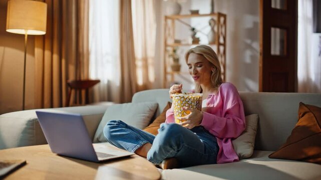 Cheerful blonde streaming series sitcom on laptop snacking chips in comfy house