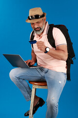 Portrait of indian middle age beard man sitting on blue studio background browsing internet on laptop shopping online holding credit card holding travel bag while sitting on the blue studio background