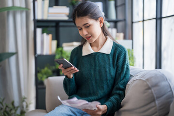 Asian woman holding paper document calculating money savings paying bills in mobile application...