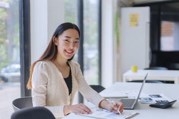 Young professional analyzing reports while working with a laptop and calculator in a bright, modern...
