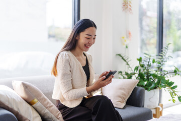 Obraz premium Young Asian businesswoman is using a smartphone and smiling while sitting on a comfortable sofa in a modern office