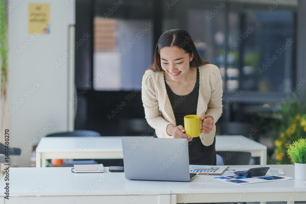 Poster young manager holding a mug and smiling while standing at her desk and using a laptop computer in a 