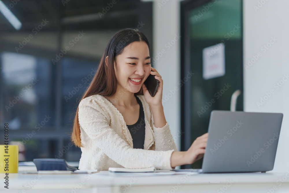 Aufkleber young office worker smiling while using a laptop and engaging in a phone call, showcasing her multit