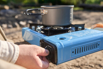 Unrecognizable traveler cooking outdoors while camping at a nature campground a sunny morning. Close up view of the hand lighting the portable stove with a pot resting on top.