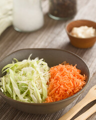 Preparing coleslaw salad made of grated carrot and chopped white cabbage in bowl, with mayonnaise, salt and black pepper in the back (Selective Focus, Focus one third into the image)