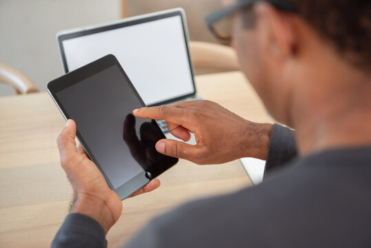 African American man using digital tablet with blank screen while sitting at desk with laptop, focusing on technology and communication in modern workspace