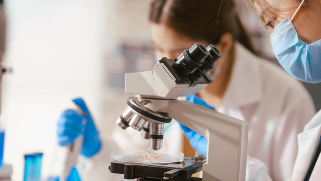 Two Asian female scientists wearing laboratory gloves sit at a table, carefully examining a test tube filled with blue liquid, using a microscope for detailed analysis in a research laboratory