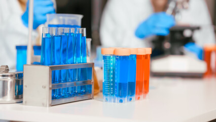 Two Asian female scientists wearing laboratory gloves sit at a table, carefully examining a test tube filled with blue liquid, using a microscope for detailed analysis in a research laboratory