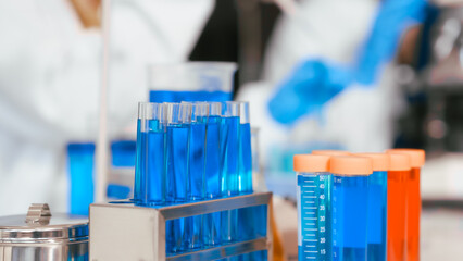 Two Asian female scientists wearing laboratory gloves sit at a table, carefully examining a test tube filled with blue liquid, using a microscope for detailed analysis in a research laboratory