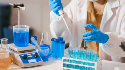 Two Asian female scientists wearing laboratory gloves sit at a table, carefully examining a test tube filled with blue liquid, using a microscope for detailed analysis in a research laboratory