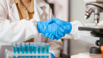 Two Asian female scientists wearing laboratory gloves sit at a table, carefully examining a test tube filled with blue liquid, using a microscope for detailed analysis in a research laboratory