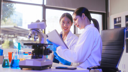 Obraz premium Two Asian female scientists wearing laboratory gloves sit at a table, carefully examining a test tube filled with blue liquid, using a microscope for detailed analysis in a research laboratory