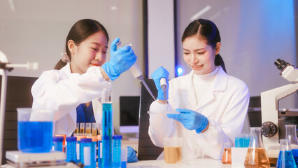 Fototapeta premium Two Asian female scientists wearing laboratory gloves sit at a table, carefully examining a test tube filled with blue liquid, using a microscope for detailed analysis in a research laboratory