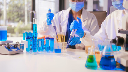 Fototapeta premium Two Asian female scientists wearing laboratory gloves sit at a table, carefully examining a test tube filled with blue liquid, using a microscope for detailed analysis in a research laboratory