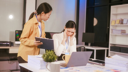 Two Asian women sit working at  desk,overwhelmed with stress struggling with headaches,feeling...