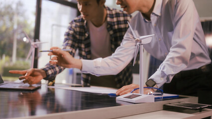 Two Asian businessmen wearing engineer hats table, discussing solar panels and wind turbines. They plan environmental projects, focusing on solar energy solutions and renewable energy installations
