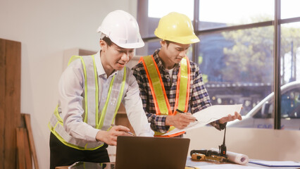 Two male engineers discuss a construction blueprint at a table in a modern office. They focus on...