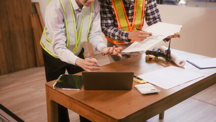 Two male engineers discuss a construction blueprint at a table in a modern office. They focus on architectural design, building structures, roofing,  wall details, planning for a contractor project