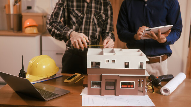 Two male engineers review blueprints in an office while discussing model house.They talk about construction projects, architectural design,roof structures,contractor coordination, building materials