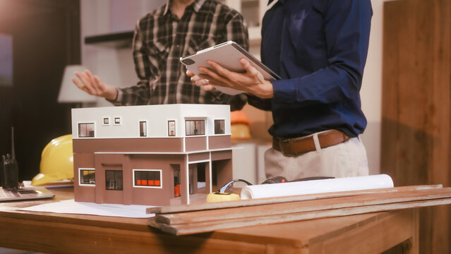 Two male engineers review blueprints in an office while discussing model house.They talk about construction projects, architectural design,roof structures,contractor coordination, building materials