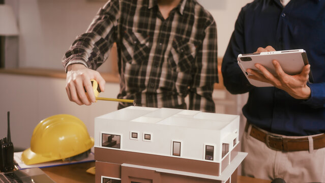Two male engineers review blueprints in an office while discussing model house.They talk about construction projects, architectural design,roof structures,contractor coordination, building materials