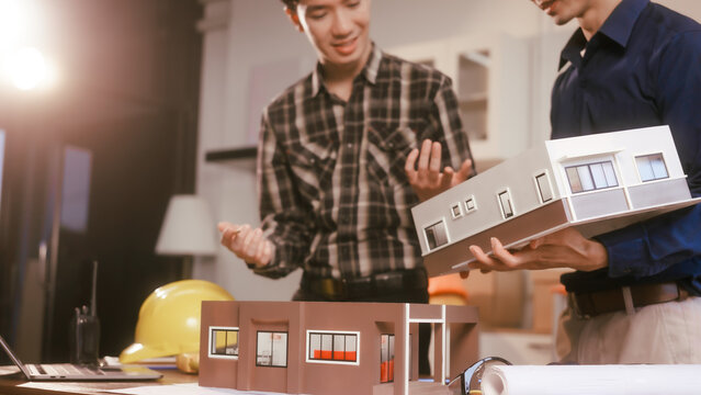 Two male engineers review blueprints in an office while discussing model house.They talk about construction projects, architectural design,roof structures,contractor coordination, building materials