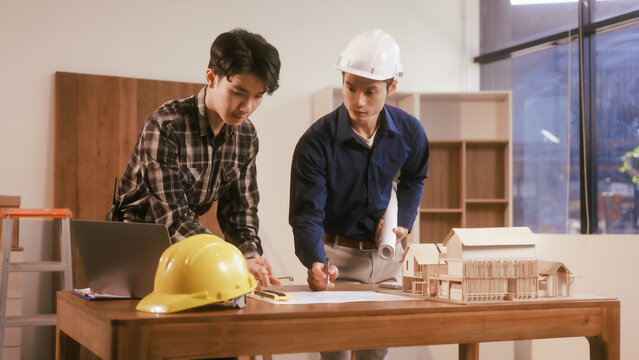 Two male engineers review blueprints in an office while discussing model house.They talk about construction projects, architectural design,roof structures,contractor coordination, building materials