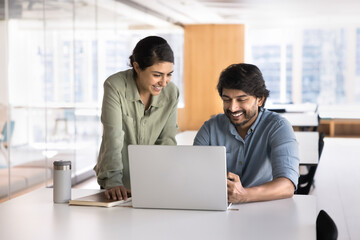 Two Indian smiling workmates working on joint task using laptop, review documents, go through...