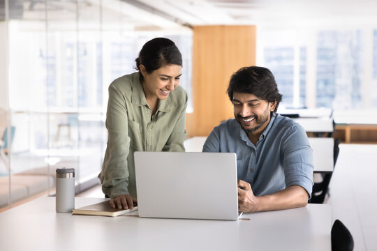 Two Indian smiling workmates working on joint task using laptop, review documents, go through reports, presentations, or proposals together, laughing, enjoy online content, brainstorm creative ideas